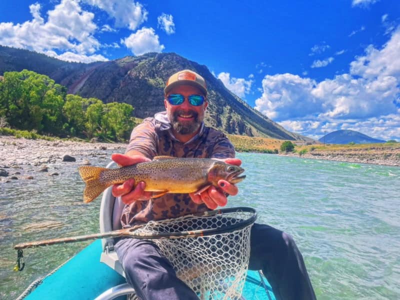Brad Pitchford in a drift boat holding a cutthroat trout, mountains and green water behind him
