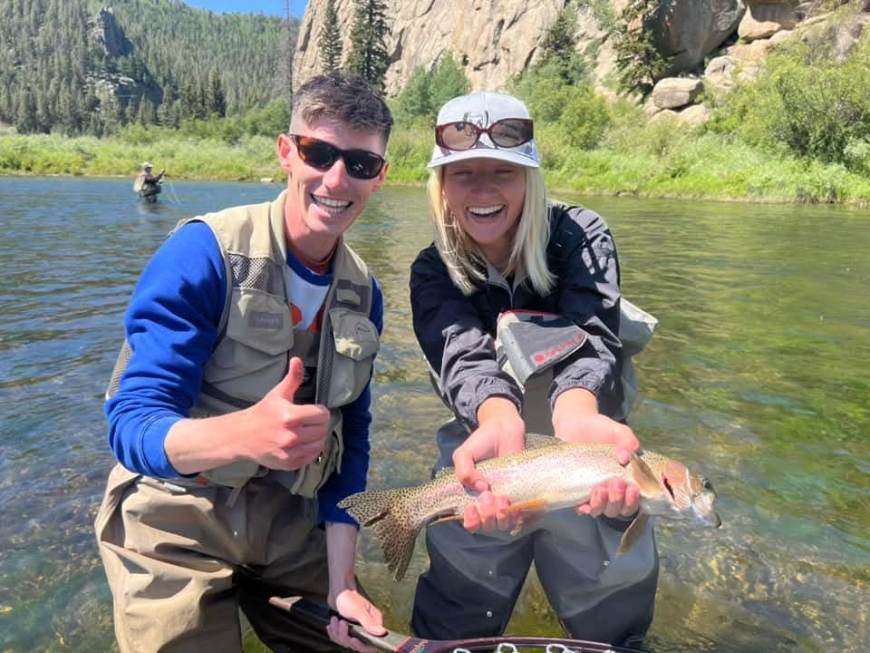 Two anglers holding a trout in a granite river canyon, with another angler wading upstream